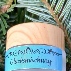 Könnte beinhalten: Ein kleines, mattiertes Glas mit einem hellen Holzdeckel. Ein hellblaues Etikett umschließt das Glas und zeigt das Wort "Glücksmischung" in einer dekorativen Schriftart. Das Glas steht vor einem Hintergrund aus grünen Tannennadeln.