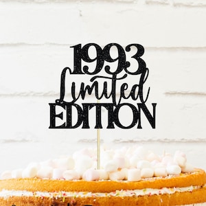 May include: Black glitter cake topper with the text "1993 Limited Edition" on a white background. The topper is on top of a yellow cake with white frosting and sprinkles.