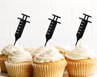 May include: Three cupcakes with black glitter syringe toppers. The cupcakes are frosted with white frosting and are on a white cake stand. The text "Etsy Shop: Cutest Little Topper" is at the bottom of the image.