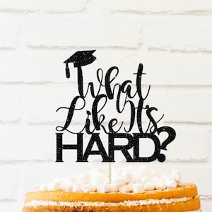 May include: Black glitter cake topper with a graduation cap and the text "What Like It's Hard?" on a white background. The topper is on top of a cake with white frosting and sprinkles.