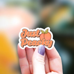 May include: A white sticker with the text "Just Peachy" in brown and orange lettering with a peach illustration. The sticker is being held by a hand.