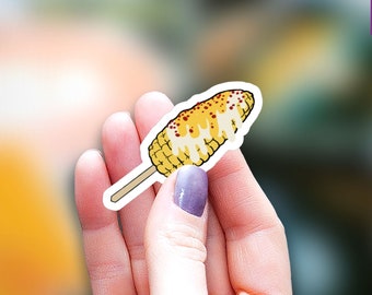 May include: A sticker of a yellow corn on the cob with white sauce and red chili flakes on top. The corn is on a stick and is held in a hand. The sticker says "Cutest Little Topper" in purple text.