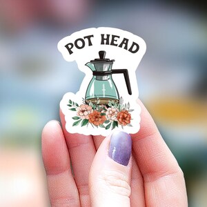 May include: A white sticker with a green glass teapot with a floral design and the text "POT HEAD" in black.