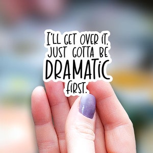 May include: A white sticker with black text that reads "I'll get over it. Just gotta be dramatic first." The sticker is held by a hand with a purple painted fingernail.