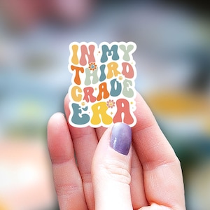 May include: A colorful sticker with the text "In My Third Grade Era" in a retro font. The sticker has a white background and features small flower designs.