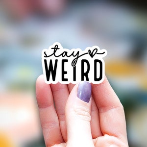 May include: White sticker with black text that reads "stay weird" with a small heart symbol above the word "weird".