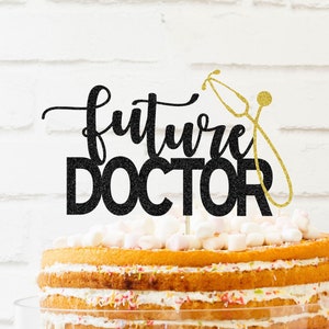 May include: Black glitter cake topper with the text "future doctor" and a gold glitter stethoscope. The topper is on a multi-layered cake with white frosting and colorful sprinkles.