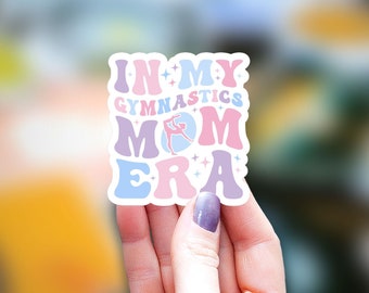 May include: A sticker with the text "In My Gymnastics Mom Era" in a retro font. The text is in pink, blue, and purple with a silhouette of a gymnast in the middle of the word "Mom".