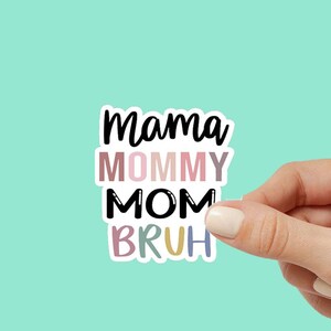 May include: A sticker with the text "Mama Mommy Mom Bruh" in black lettering with a colorful background. The sticker is being held by a hand.