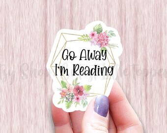 May include: A white sticker with a gold geometric frame and pink watercolor flowers. The sticker says "Go Away I'm Reading".