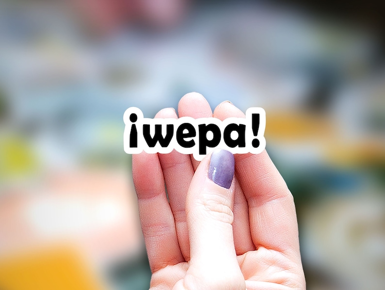 May include: A hand holding a white sticker with the text "&iexcl;wepa!" in black letters.