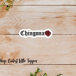 Pu&ograve; includere: Un adesivo bianco con testo nero che recita "Chingona" e una grafica di rosa rossa. L'adesivo &egrave; su uno sfondo di legno.
