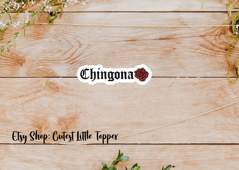 Pu&ograve; includere: Un adesivo bianco con testo nero che recita "Chingona" e una grafica di rosa rossa. L'adesivo &egrave; su uno sfondo di legno.