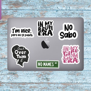 May include: A silver laptop with six stickers on it. The stickers are black and pink with various phrases, including "I'm nice, pero no te pases", "In my bruh era", "No Sabo", "Don't over think it", "No Mames", and "In my girl mom era".