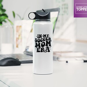 May include: White stainless steel water bottle with a black lid and a black handle. The bottle has a black graphic that says "In My Soccer Mom Era" with a football.