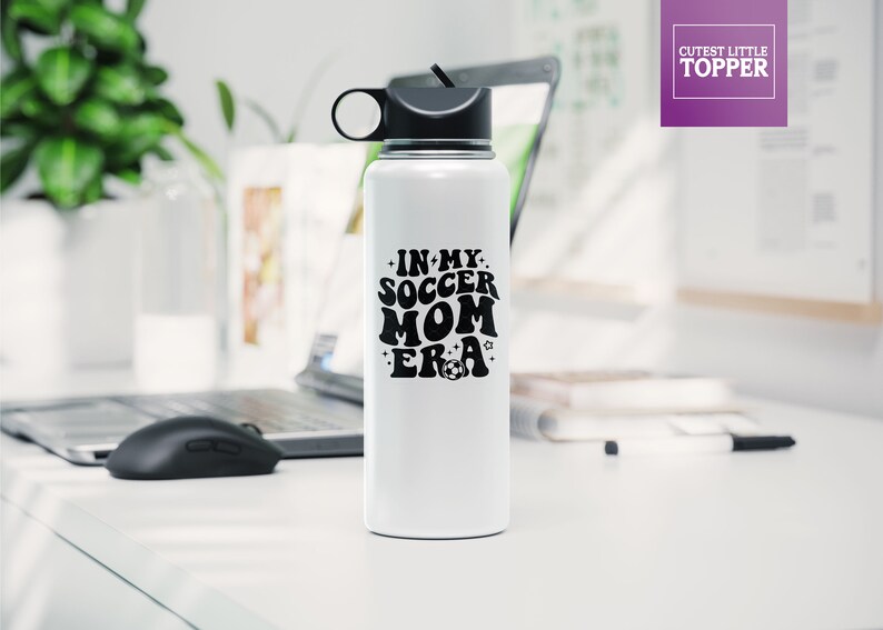 May include: White stainless steel water bottle with a black lid and a black handle. The bottle has a black graphic that says "In My Soccer Mom Era" with a football.