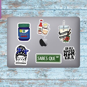 K&ouml;nnte beinhalten: Ein Laptop mit sechs Aufklebern. Die Aufkleber zeigen eine Frau mit einem Dutt und Fu&szlig;ball-Sonnenbrillen, ein Stra&szlig;enschild mit der Aufschrift "Sabes Que", eine Flasche Medizin, eine Tasse Horchata, eine Tasse Instantnudeln und einen Aufkleber mit der Aufschrift "In My Soccer Mom Era".