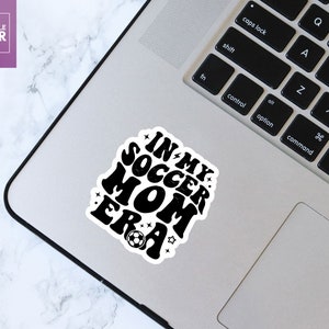 May include: Black and white sticker with the text "In My Soccer Mom Era" in a retro font. A football is below the text.