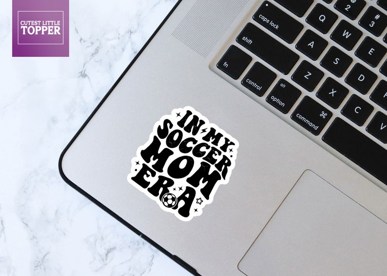 May include: Black and white sticker with the text "In My Soccer Mom Era" in a retro font. A football is below the text.