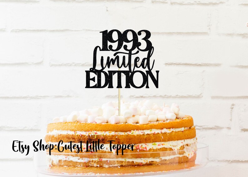 May include: Black glitter cake topper with the text "1993 Limited Edition" on a white background. The topper is on top of a yellow cake with white frosting and sprinkles.
