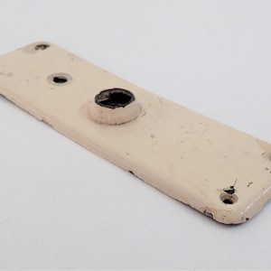 Könnte beinhalten: Rechteckige, cremefarbene Metalltürplatte mit erhabenem, kreisförmigem Abschnitt und vier Schraubenlöchern. Die Farbe ist abgeblättert und gibt den Blick auf das Metall frei. Antikes Objekt.
