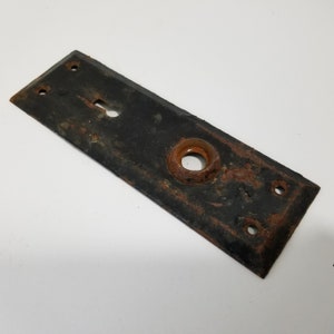 Puede incluir: Una placa de puerta negra rectangular antigua con un ojo de cerradura y una abertura circular para un pomo. La placa de metal muestra signos de óxido y desgaste, con cuatro agujeros para tornillos.