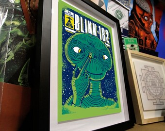 Può includere: Una stampa artistica incorniciata raffigurante un personaggio alieno verde che si tocca il naso, con il testo "BLINK-182" e "WITH STATE CHAMPS" in alto. L'illustrazione è su uno sfondo blu stellato, in una cornice nera con passe-partout bianco.