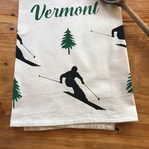 May include: A white kitchen towel with the word "Vermont" printed in green cursive script. The towel features a repeating pattern of green pine trees and black silhouettes of skiers.