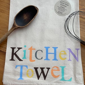 Puede incluir: Una toalla de cocina de saco de harina blanca con las palabras "Kitchen Towel" impresas en letras de colores. La toalla está hecha de 100% algodón natural sin blanquear y es lavable a máquina.