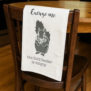 May include: A white kitchen towel with a squirrel graphic and the text "Excuse me the bird feeder is empty" is draped over a wooden chair. The squirrel is gray and the text is black.