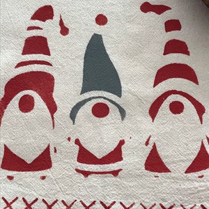 May include: A white kitchen towel with a red and gray gnome design. The gnomes are wearing red hats with white stripes and have red noses. The towel has a red stitched border with an 'X' pattern.