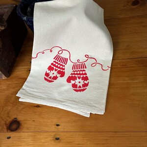 Puede incluir: Una toalla de cocina blanca doblada con un diseño de mitones rojos. Los mitones están conectados por una línea roja y presentan un patrón de copo de nieve. La toalla está hecha de un material suave y absorbente.