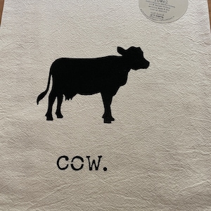 Puede incluir: Una toalla de cocina de saco de harina blanca con una silueta negra de una vaca y la palabra "COW." impresa en ella. La toalla está hecha de 100% algodón y está impresa a mano individualmente.