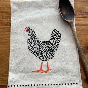 Puede incluir: Una toalla de cocina blanca con una ilustración en blanco y negro de una gallina. La gallina tiene patas naranjas y una cresta roja. La toalla tiene un borde de puntos negros.