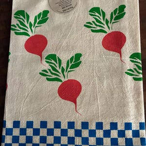 Puede incluir: Un paño de cocina de color crema con un patrón repetitivo de rábanos rojos con hojas verdes. Un borde a cuadros azules y blancos recorre el borde inferior. Una etiqueta circular en la parte superior dice Flour Sack Kitchen Towel.