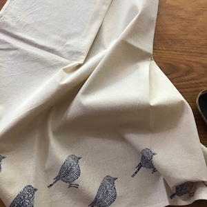 Puede incluir: Un pa&ntilde;o de cocina de algod&oacute;n blanco con un dise&ntilde;o de p&aacute;jaros azules. Los p&aacute;jaros est&aacute;n impresos en un estilo de impresi&oacute;n en bloque y est&aacute;n dispersos por el pa&ntilde;o.