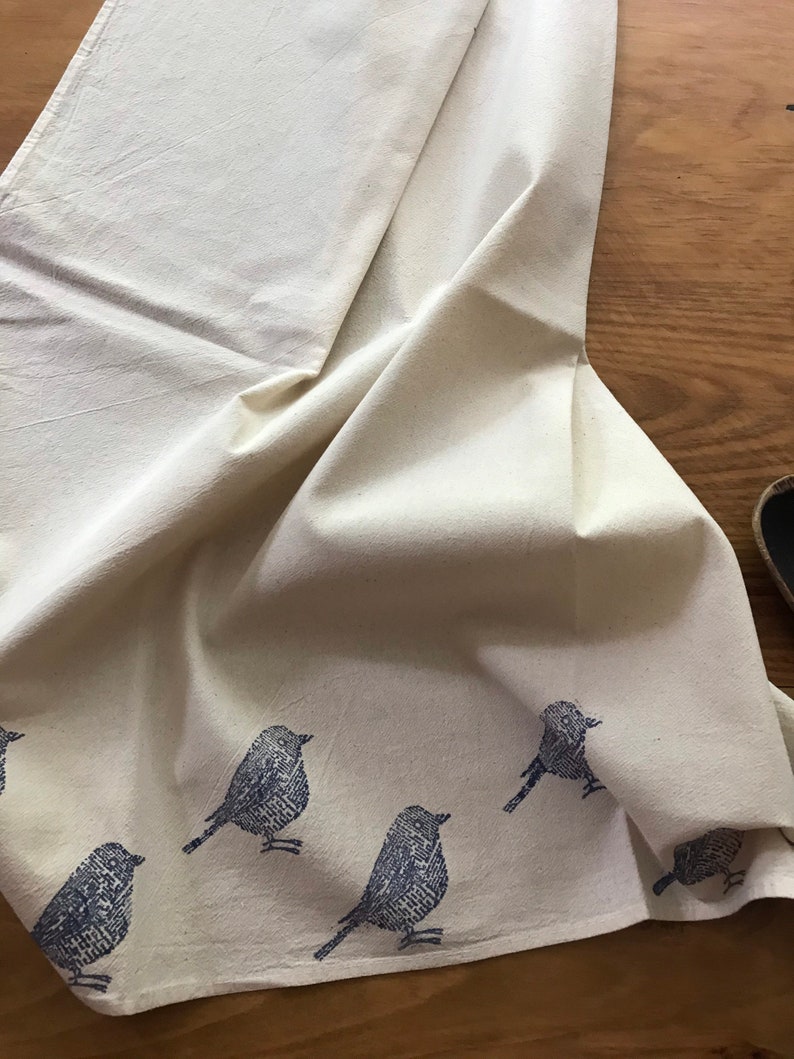 Puede incluir: Un pa&ntilde;o de cocina de algod&oacute;n blanco con un dise&ntilde;o de p&aacute;jaros azules. Los p&aacute;jaros est&aacute;n impresos en un estilo de impresi&oacute;n en bloque y est&aacute;n dispersos por el pa&ntilde;o.