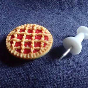 May include: A small pie with a lattice design and red filling is next to a white drawing pin with a silver pin. The pie has a golden-brown crust. The drawing pin is on a blue surface.