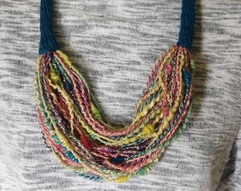 May include: A colorful yarn necklace with a teal cord. The necklace is made of multiple strands of yarn in various shades of pink, yellow, green, and blue.