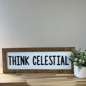May include: A wooden framed sign with the text "THINK CELESTIAL" in black letters on a white background.