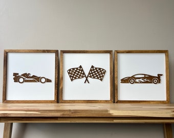May include: Three framed wooden wall art pieces featuring racing themes. One depicts a race car, another crossed checkered flags, and the third a sports car with flames. Each piece has a white background and a natural wood frame.