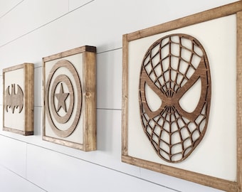 May include: Three framed wooden wall art pieces featuring superhero symbols. The first displays the Batman logo, the second Captain America's shield, and the third Spider-Man's mask. Each piece has a light-colored background and a dark wood frame.