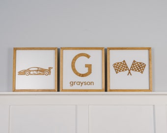 May include: Three framed wall art pieces with a racing theme. One features a race car with flames, another a letter "G" with the name "grayson" below, and the third shows crossed checkered flags. All are in wooden frames.