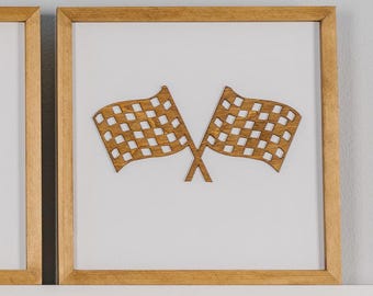 May include: Two wooden checkered racing flags in a square, light-brown framed artwork. The flags are a light brown color with a checkered pattern. The artwork is set against a white background.