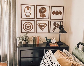 May include: Six framed wooden wall art pieces featuring superhero symbols: Iron Man's mask, a fist, Batman's logo, Captain America's shield, Superman's emblem, and Thor's hammer. The art is displayed above a black dresser.