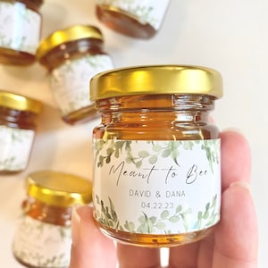 May include: Small glass jars filled with honey, with a white label that reads "Meant to Bee" with a floral design and the text "David & Dana 04.22.23".