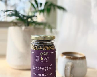 May include: A glass jar of loose leaf tea labeled "Oil of Joy Apothecary Decongest Organic Tea Blend" with a purple label and gold lid.