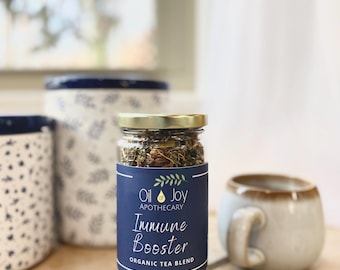 May include: A glass jar filled with dried herbs and flowers. The jar has a blue label with white text that reads "Oil of Joy Apothecary Immune Booster Organic Tea Blend".