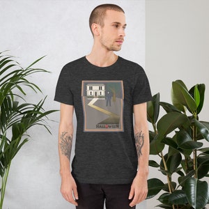 Könnte beinhalten: Ein dunkel-helles graues T-Shirt mit einem Grafikdruck eines Hauses und einer Figur davor. Die Grafik ist in einem braunen Rahmen mit dem Wort "HALLOWEEN" darunter.