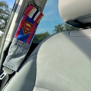May include: A gray and blue car seat belt cover with a Superman logo and a skull and crossbones design. The cover is attached to a car seat belt.